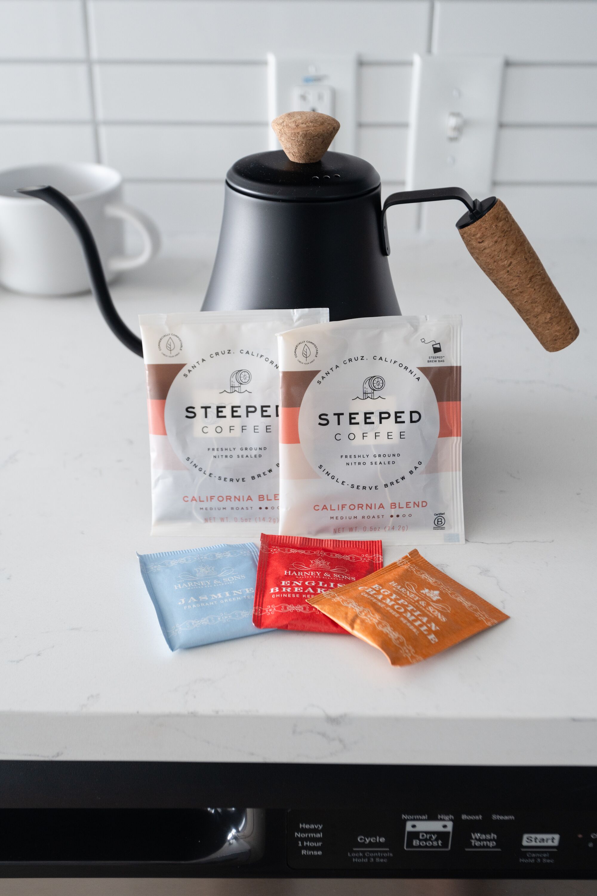 Une machine à café dotée d’un bouchon en liège, deux sachets de café infusé et trois sachets de thé Harney & Sons, le tout posé sur un plan de travail de cuisine.