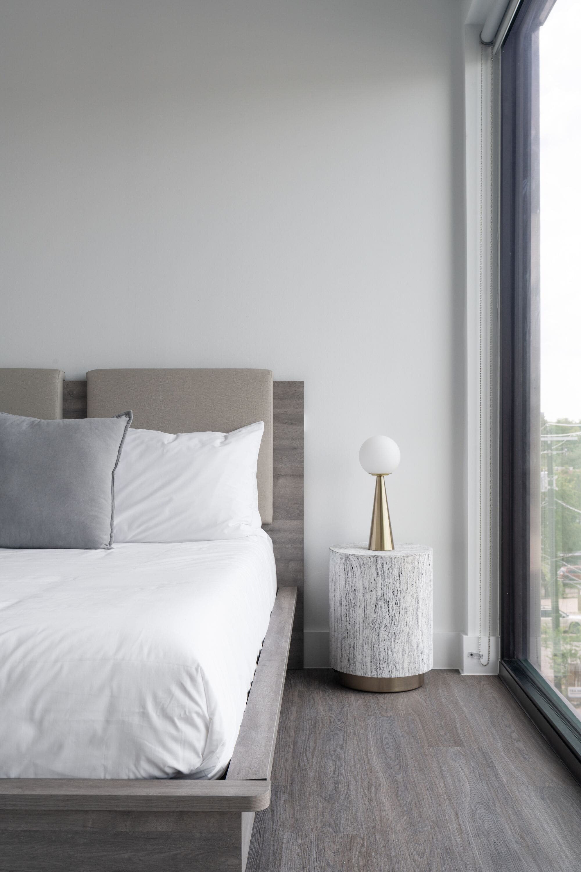 A modern bedroom with a large bed, a round nightstand, and a view through a window.