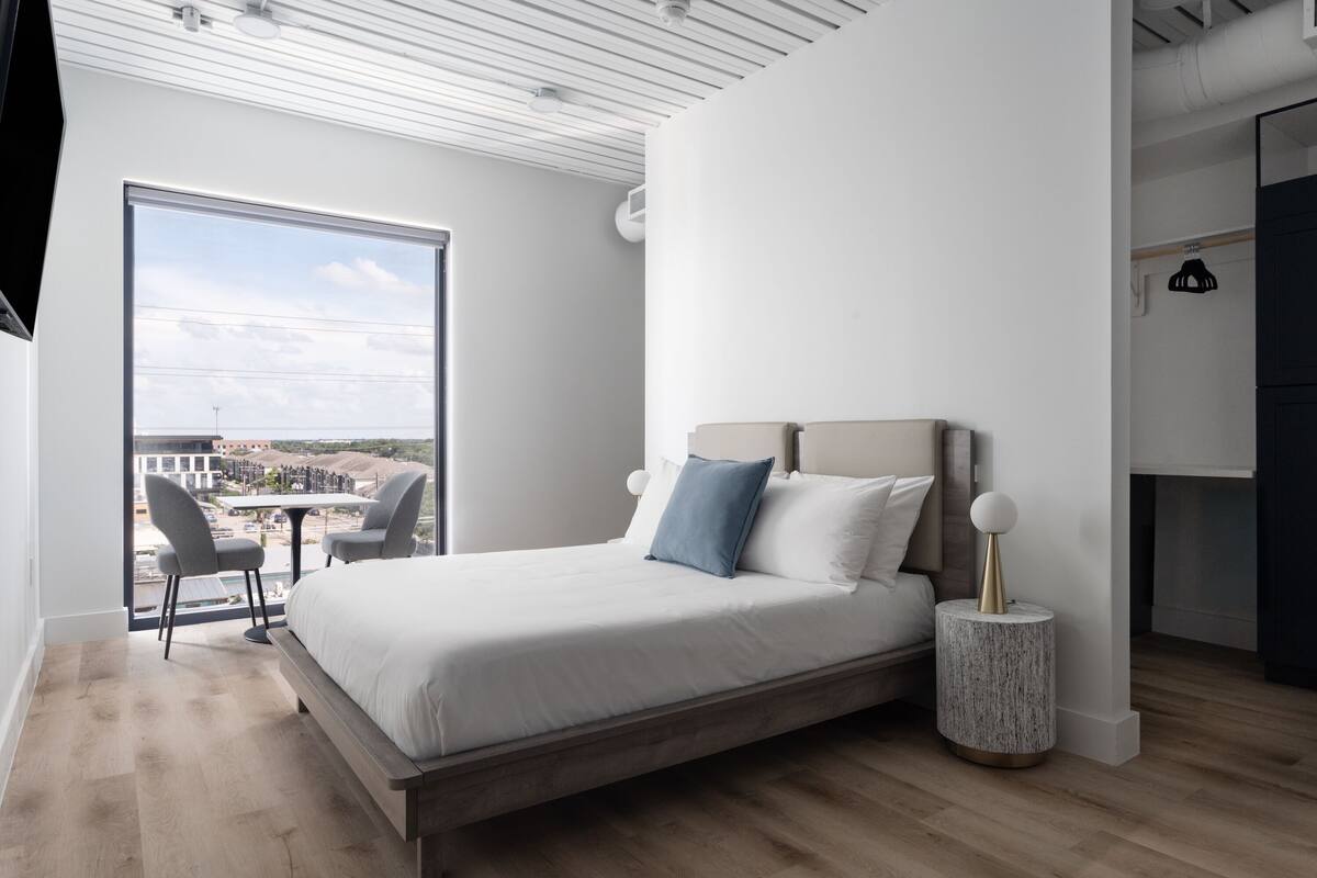 Un dormitorio moderno con una cama grande, una mesita redonda y vistas de la ciudad a través de un ventanal amplio.