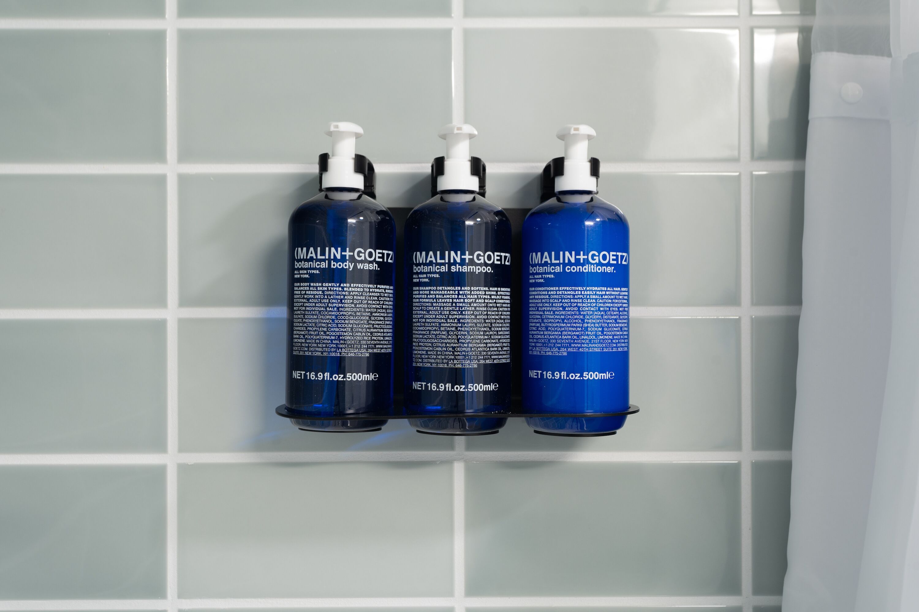 Three bottles of MALIN+GOETZ botanical body wash, shampoo, and conditioner on a tiled wall.