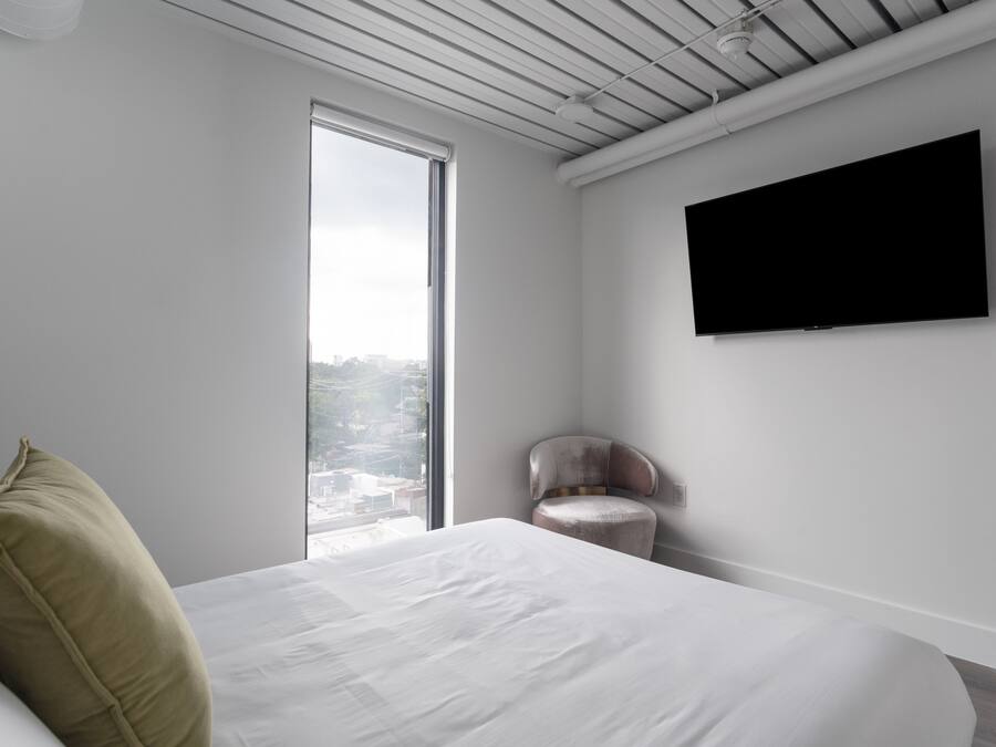 A modern hotel room with a large bed, a flat-screen TV, a chair, and a city view.