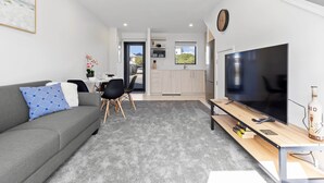 A modern living room with a grey sofa, a coffee table, and a TV. There is a dining area with a table and chairs, and a kitchen with cabinets and a window. - Beachside Townhouse - New Brighton (Christchurch)