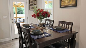 Comedor con mesa de madera dispuesta para cuatro personas, rodeada de sillas. Hay un jarrón con flores rojas sobre la mesa, y un cuadro enmarcado en la pared.