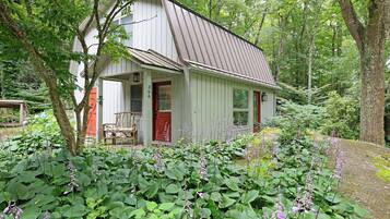 A white wooden house with a red door, surrounded by lush greenery and flowering plants.