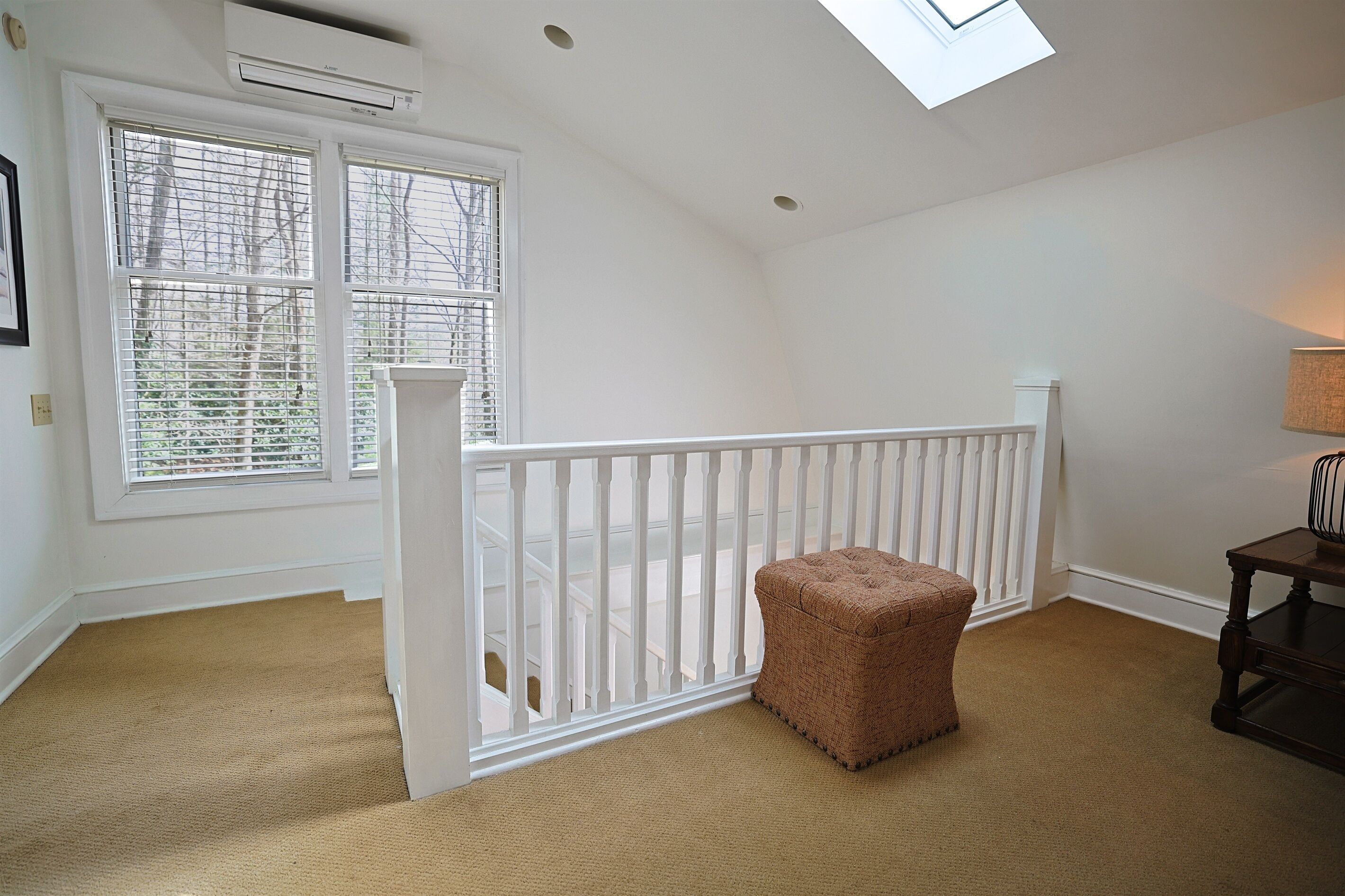 A room with a white railing, a brown ottoman, and a window with blinds.