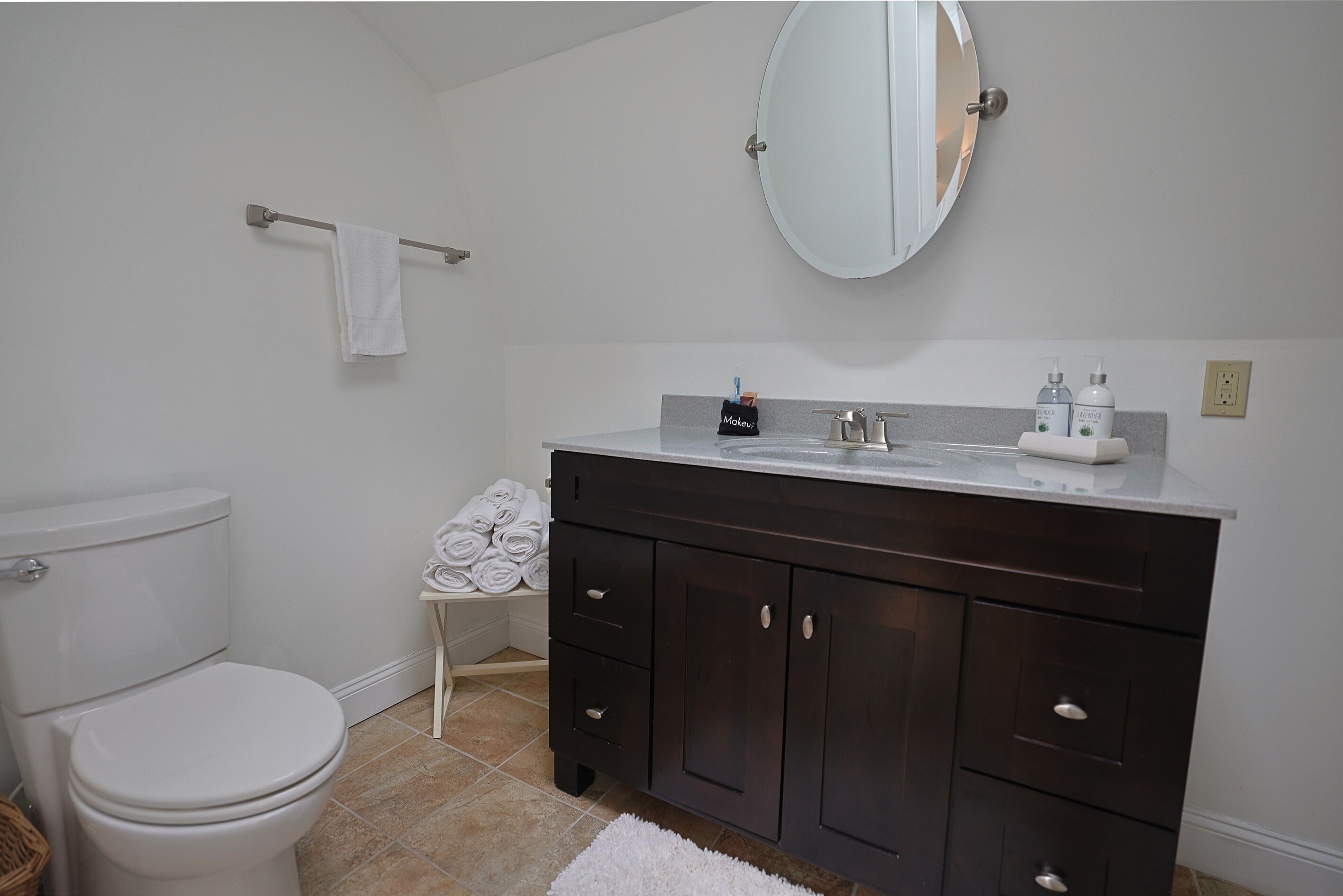 A bathroom with a white toilet, dark wooden vanity, a round mirror, and a towel rack.