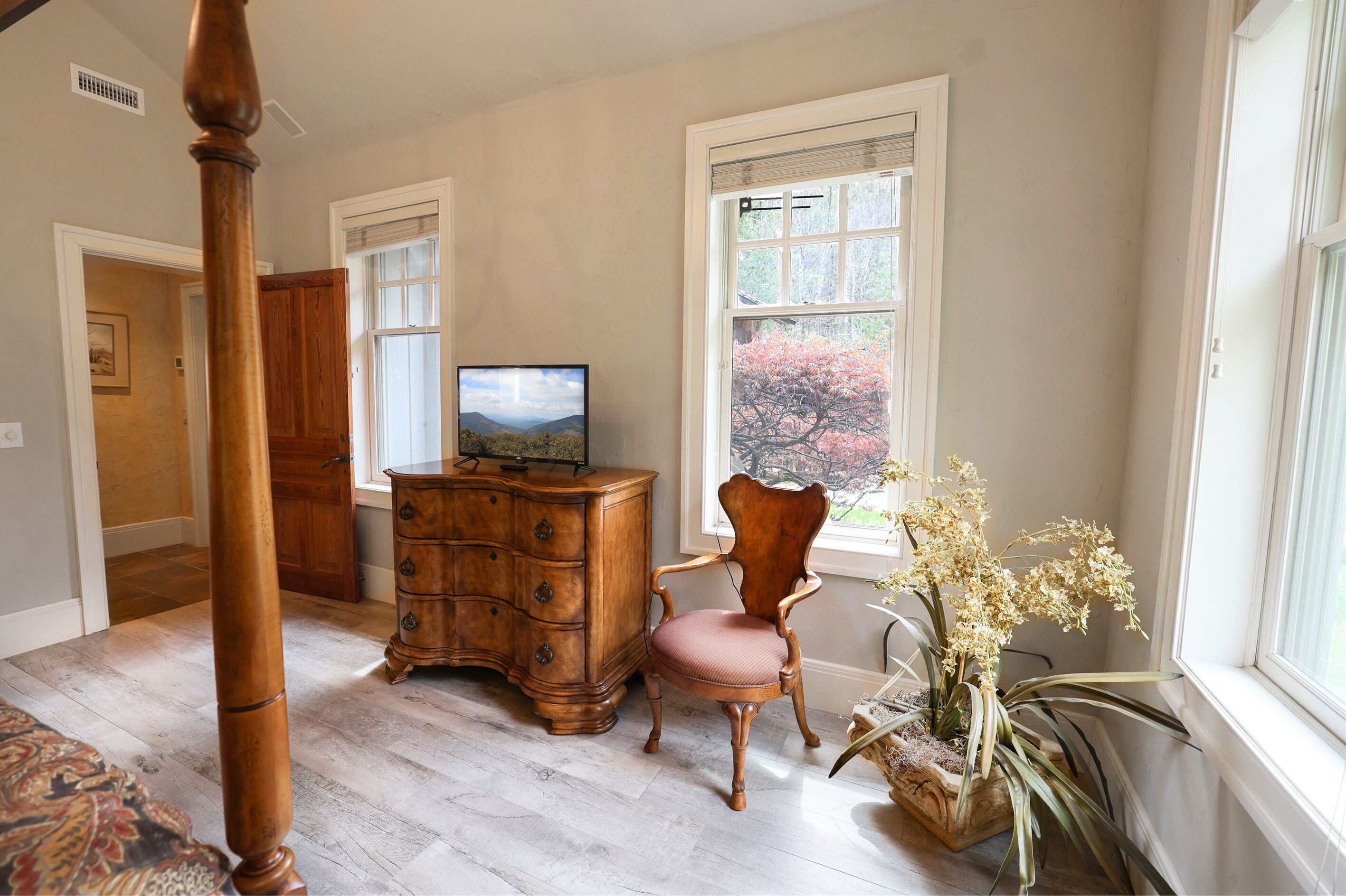 A room with a wooden dresser, a chair, a television, and a window with a view of trees.