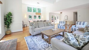 A spacious living room with a grey sofa, blue patterned armchairs, a wooden coffee table, and a large window providing natural light. - Lg Pool House w/ Hot Tub Central to RTP*Pool OPEN* (Durham)