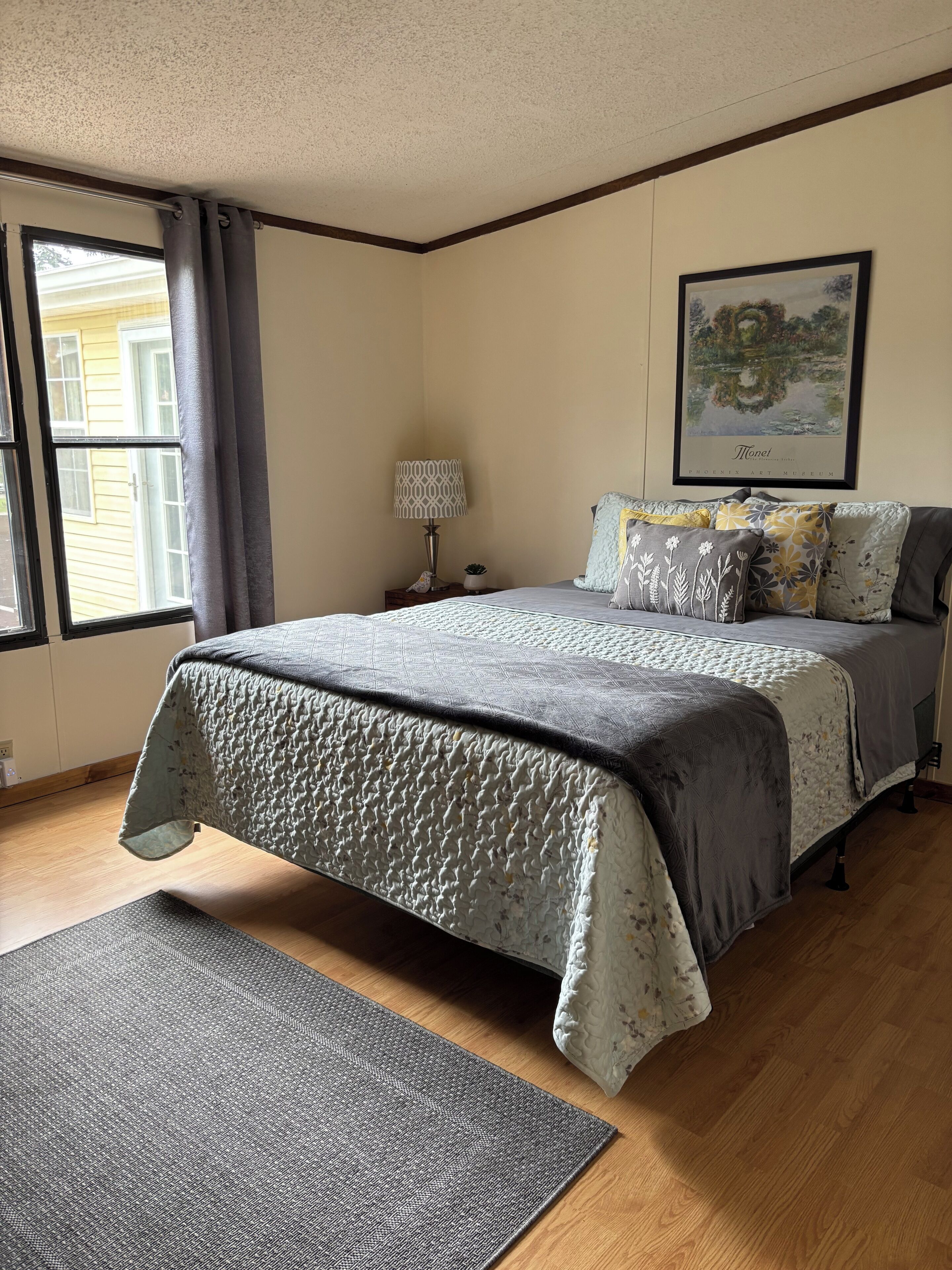 Une chambre à coucher avec un lit, une table de chevet, une lampe, une fenêtre avec des rideaux et un tableau encadré au mur.