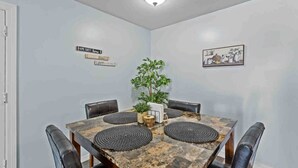 Une salle à manger avec une table carrée, des chaises en cuir noir, une plante en pot et des panneaux décoratifs fixés au mur.