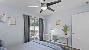 Une chambre à coucher comprenant un lit, un bureau, une chaise, un ventilateur de plafond, une fenêtre avec des rideaux et une porte.