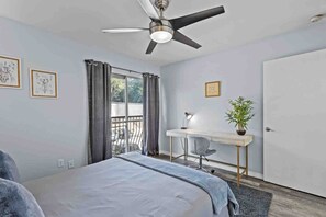 Een slaapkamer met een bed, een bureau, een stoel, een plafondventilator, een raam met gordijnen en een deur.