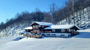 Een ruime houten chalet met meerdere balkons, gelegen op een met sneeuw bedekte helling met een bos op de achtergrond.