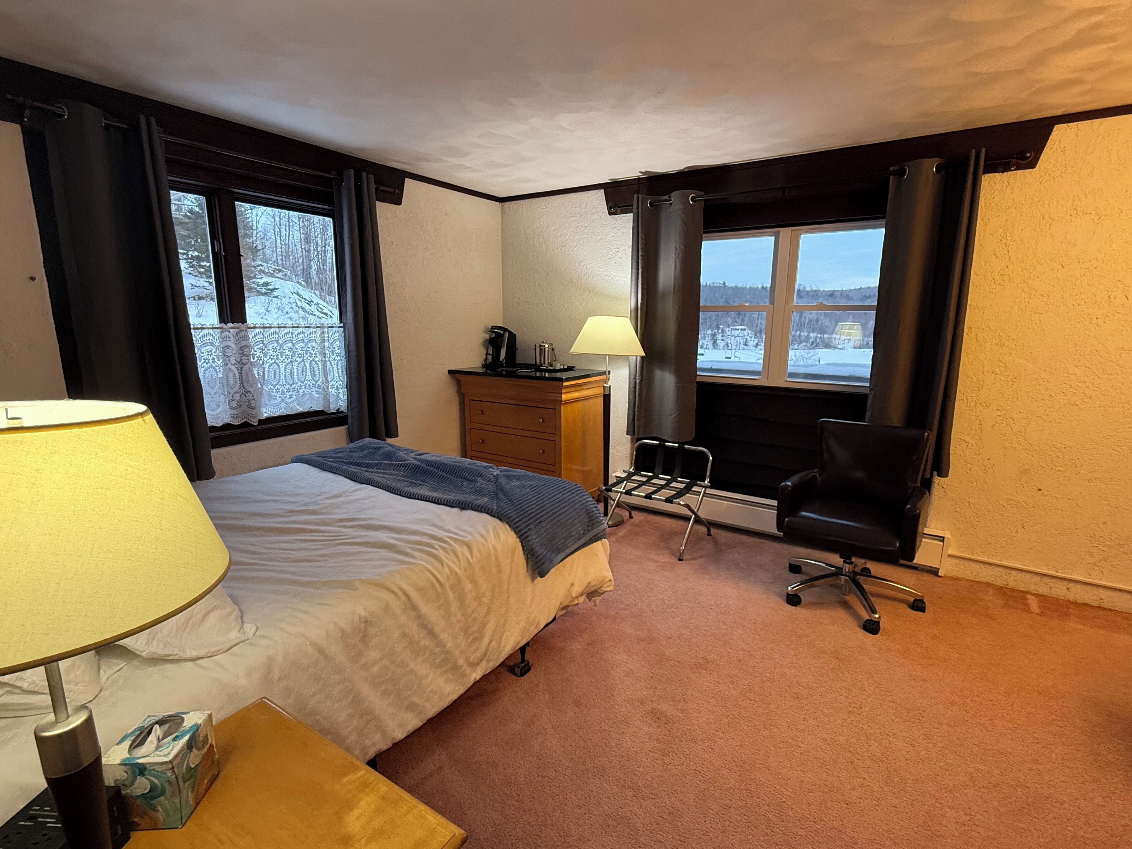 Habitación de hotel con cama, escritorio, silla, lámpara y vista a un paisaje nevado a través de las ventanas.