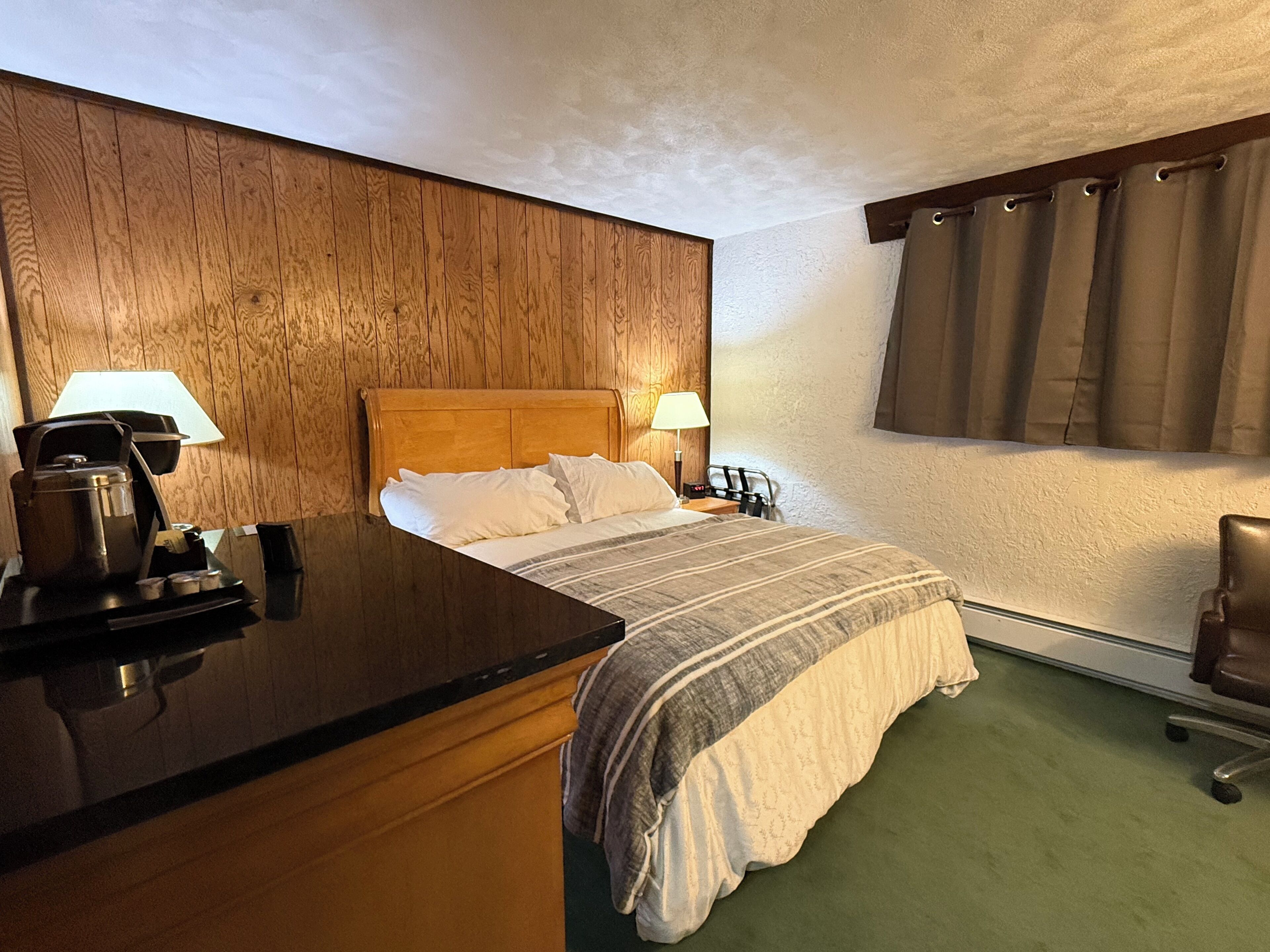 Ett hotellrum med en sänggavel i trä, en säng med vita sängkläder, ett nattduksbord med en lampa och en grön matta.