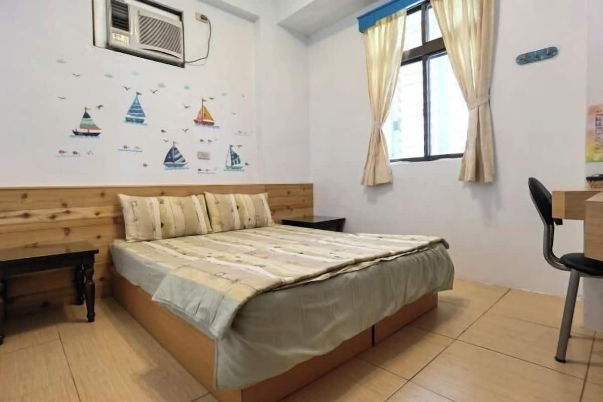 Quarto com cabeceira de madeira, cama com colcha listrada, escrivaninha com cadeira, janela com cortinas e parede com adesivos de barcos à vela.