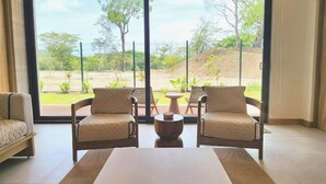 A room with a large sliding glass door leading to an outdoor area with greenery and a view of a hill. Inside, there is a sofa, a coffee table, and a chair. - C14 - les Domus

 (Nicoya)