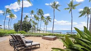 A beachside lounge area with lounge chairs and a small wooden table, surrounded by palm trees and a view of the ocean. - Highly Rated 7th Floor End Unit with Amazing Lanai Views. Book Ahead & Save!! (Kapolei)