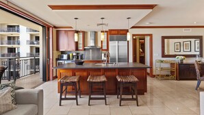 A spacious kitchen with a large island, wooden cabinets, and a stainless steel refrigerator. There are three bar stools in front of the island. - Highly Rated 7th Floor End Unit with Amazing Lanai Views. Book Ahead & Save!! (Kapolei)