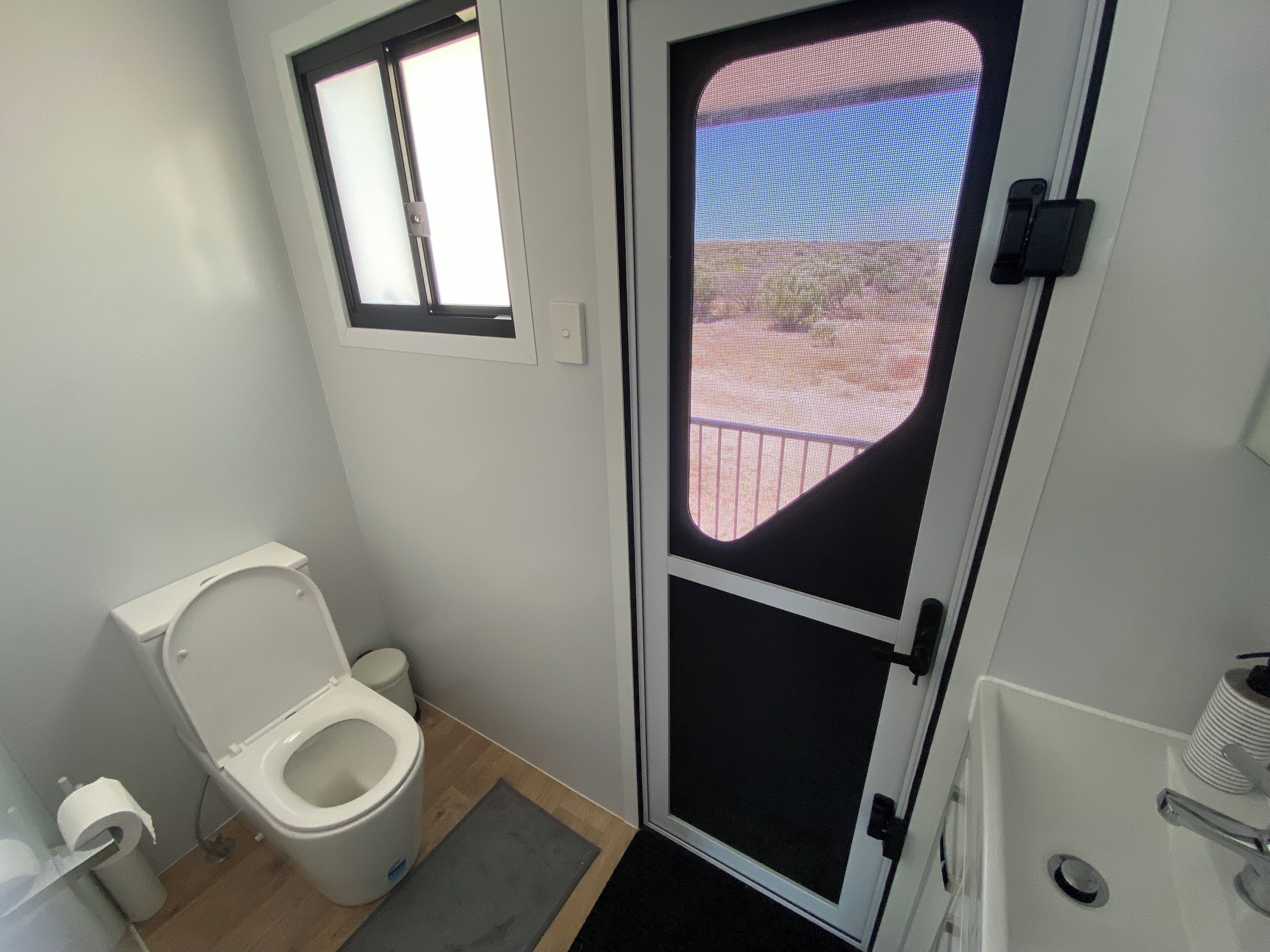 Een compacte badkamer met toilet, een klein raam en een deur met uitzicht op buiten.