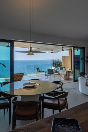 A modern dining area with a view of the sea, featuring a round table, wooden chairs, and a glass partition. - Villa Cocó (Punta Mita)