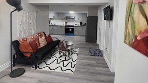 A modern living room with a sofa, a coffee table, and a floor lamp. There is a kitchen in the background with cabinets and a refrigerator.