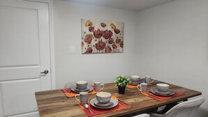 A dining area with a wooden table set for four, surrounded by grey chairs. A floral painting hangs on the wall above the table.