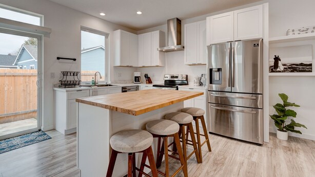A modern kitchen with white cabinets, a stainless steel refrigerator, a wooden island with stools, and a large window. - Brand New Family-Friendly Home for Rent in Battle Ground, WA (Battle Ground)