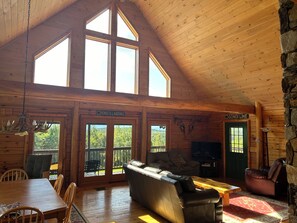 A log cabin interior with a dining area, a sofa, a television, and a stone wall. - High View Lodge, log chalet with unparalleled views of Western Maine Mountains (Eustis)