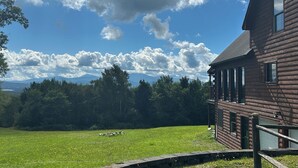 Een houten hut met een veranda, omgeven door een grasveld en bomen, met bergen op de achtergrond.