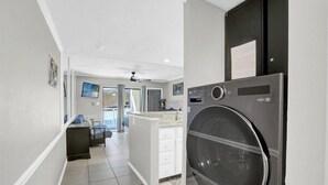 A laundry room with a large front-loading washer and dryer, a small kitchenette with a countertop and sink, and a hallway leading to a bedroom with a bed and a desk.