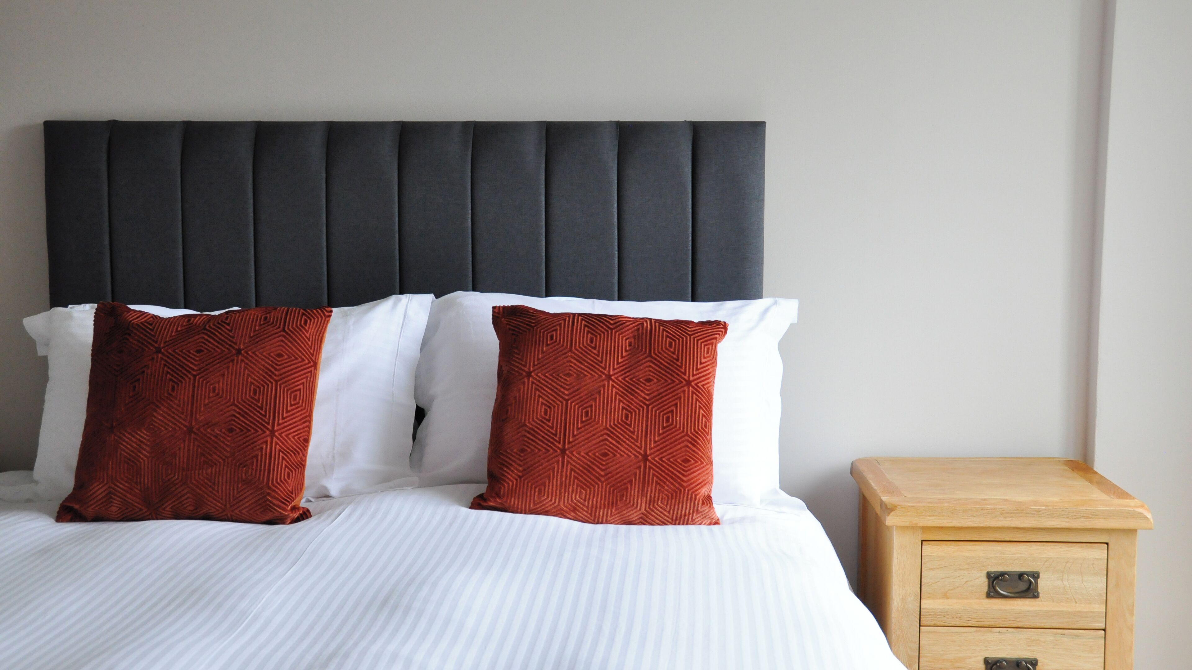 Un lit avec une literie blanche et deux coussins rouges à motifs, une tête de lit sombre recouverte de tissu et une table de chevet en bois avec des tiroirs.