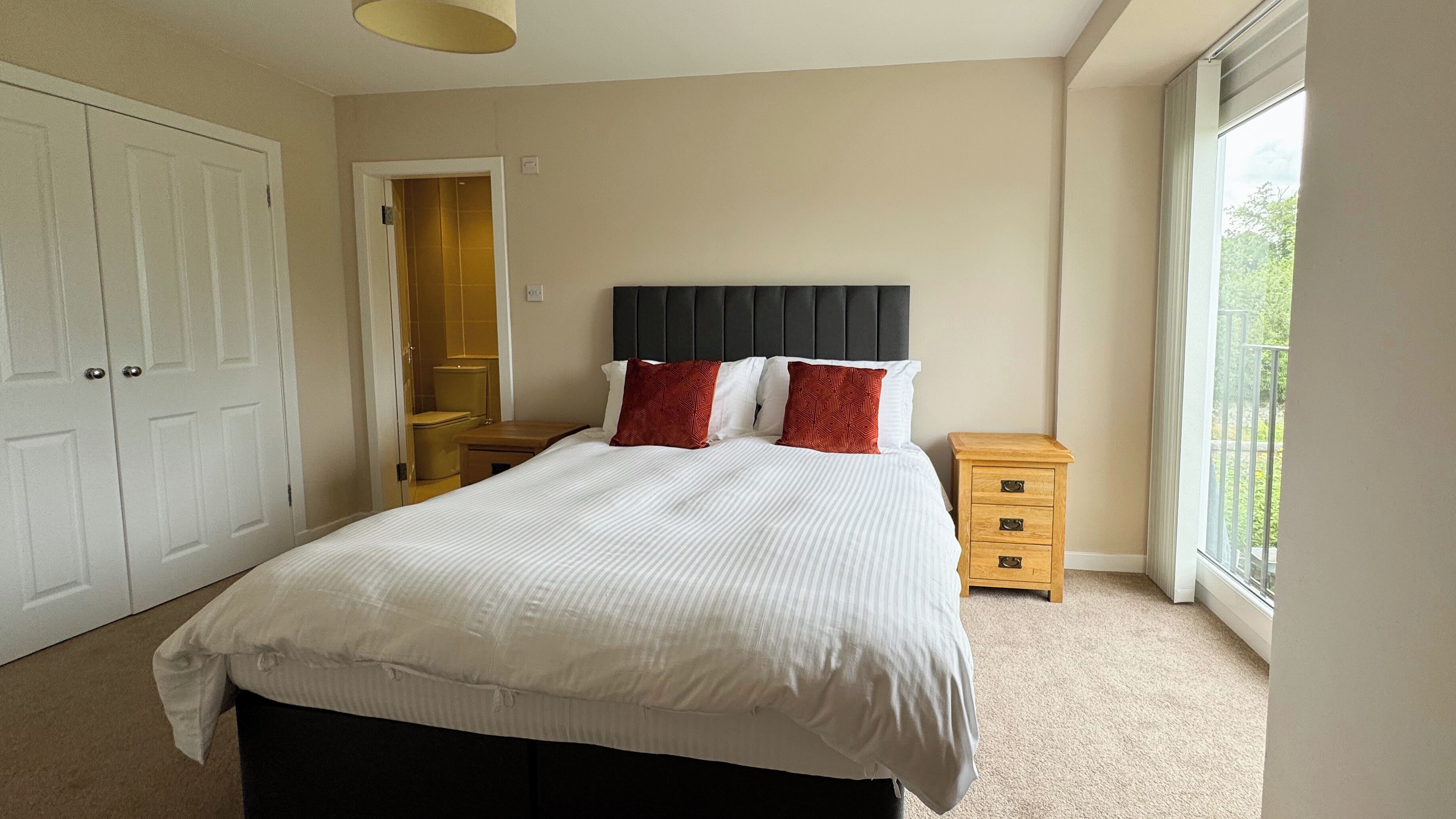A bedroom with a bed, a nightstand, a wardrobe, and a bathroom visible through an open door.