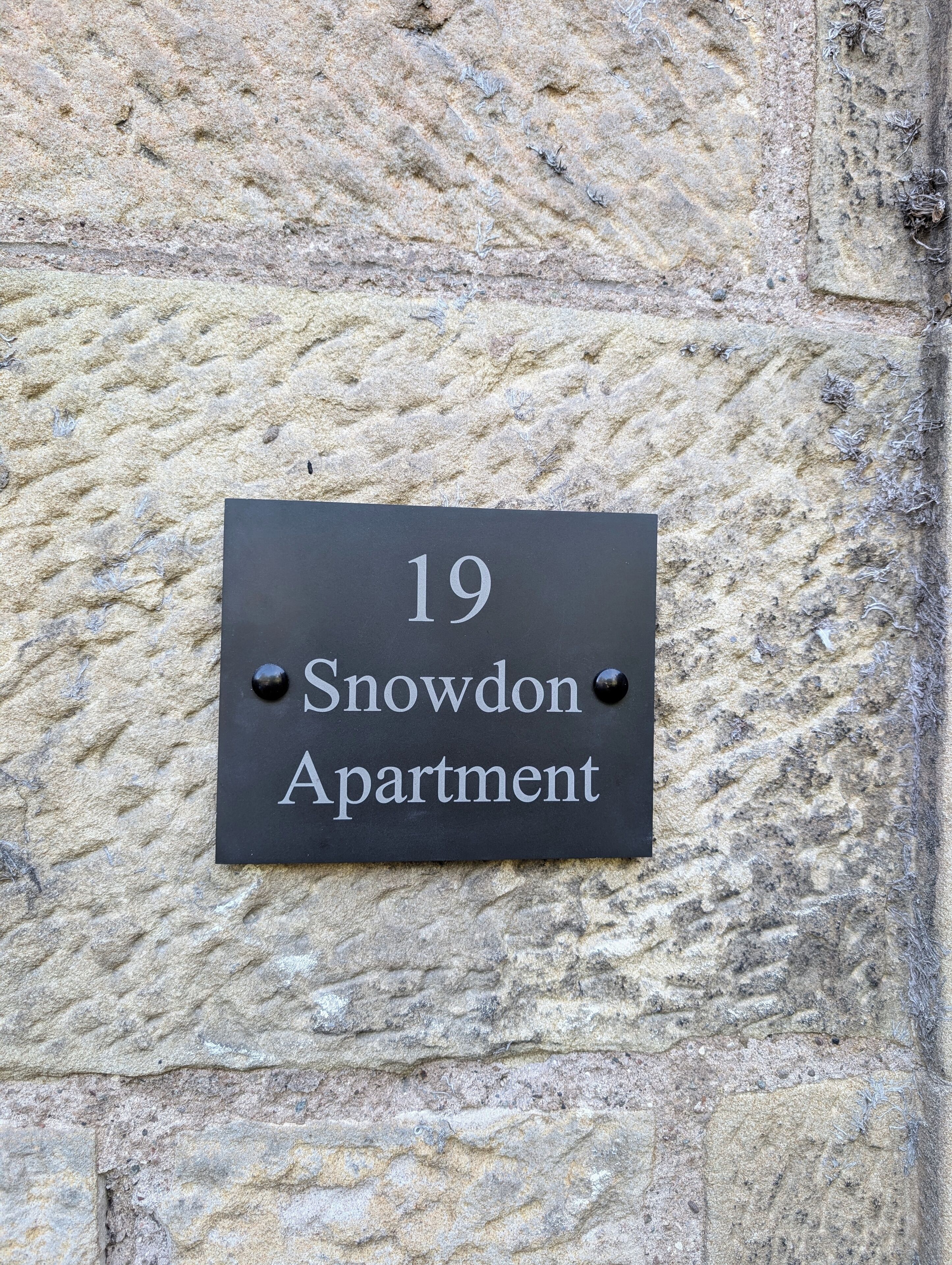 Une plaque noire avec une inscription blanche sur un mur de pierre à la texture particulière, portant l’inscription « Appartement 19, Snowdon ».