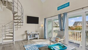 Ein Wohnzimmer mit Wendeltreppe, einem Flachbildfernseher, einem kleinen Tisch mit Tablett und Blick auf einen Balkon mit dem Schriftzug "EMERALD ISLE".