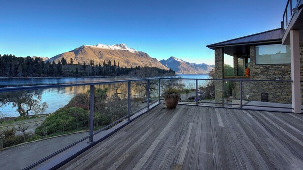 Une terrasse en bois dotée d’une balustrade en verre, offrant une vue sur un lac et des montagnes.