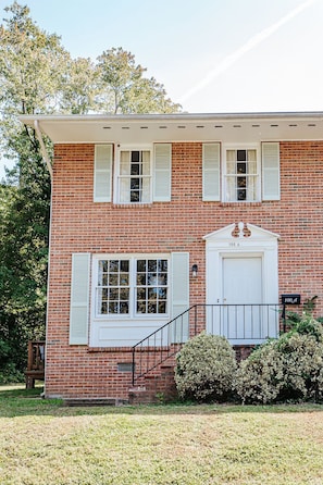 A two-story brick building with a white front door, two windows with shutters, a small porch, and a mailbox.
