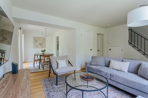 A modern living room with a grey sofa, a round glass coffee table, a wooden side table, a dining area with a table and chairs, and a staircase with a railing.