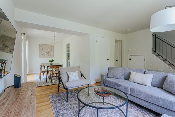 A modern living room with a grey sofa, a round glass coffee table, a wooden side table, a dining area with a table and chairs, and a staircase with a railing.