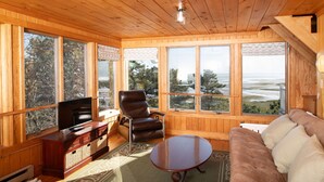 A wooden-paneled room with a television, leather chair, sofa, and large windows offering a view of trees and water. - Family & Pet Getaway | Scenic Views |Cape Cod Home (Wellfleet)