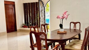 Uma sala de jantar com uma mesa de madeira redonda, seis cadeiras de madeira com assentos acolchoados e um vaso de plantas com flores cor-de-rosa.