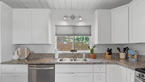 Cocina con armarios blancos, encimeras de granito, lavavajillas, ventana con persianas y una planta en maceta.