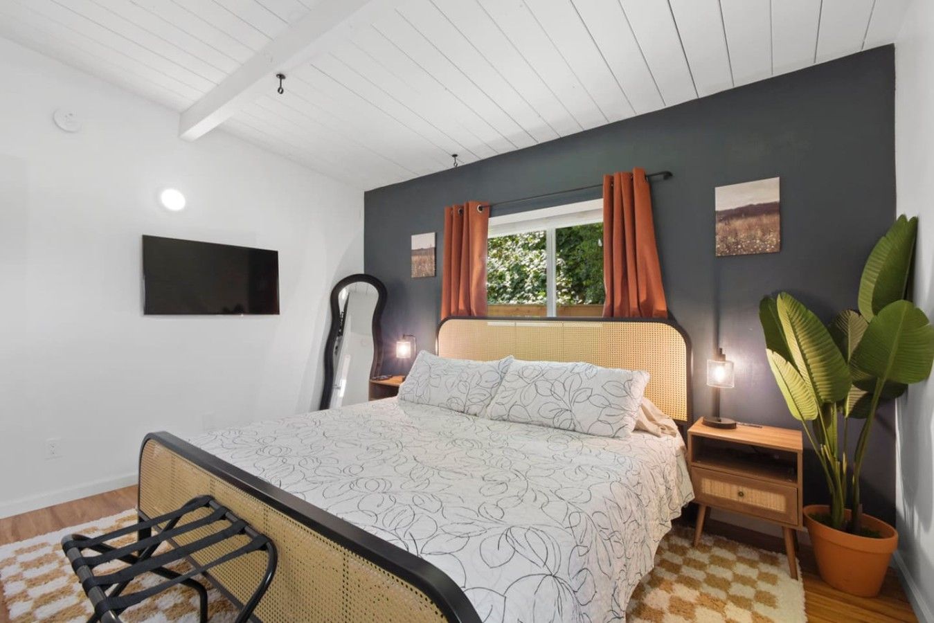 A bedroom with a bed, a bedside table, a lamp, a mirror, a potted plant, and a window with curtains.