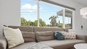 Una sala de estar moderna con un sofá color beige, un plaid estampado y vistas a árboles y un cielo despejado a través de amplios ventanales.