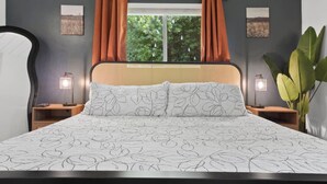 Un dormitorio con una cama, dos mesitas de noche, un ventanal con cortinas y dos lámparas de pared.