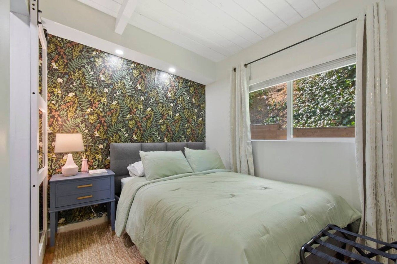 Un dormitorio con papel tapiz floral, una cama con ropa de cama verde, una mesita de noche con lámpara y una ventana con cortinas.