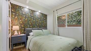 Un dormitorio con papel tapiz floral, una cama con ropa de cama verde, una mesita de noche con lámpara y una ventana con cortinas.