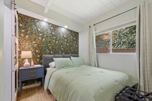 A bedroom with a floral wallpaper, a bed with green bedding, a nightstand with a lamp, and a window with curtains.