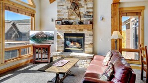 Una sala de estar acogedora con chimenea de piedra, vigas de madera y un amplio ventanal con vistas a un paisaje montañoso.