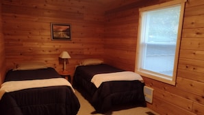Une chambre de style cabane avec deux lits, une table de chevet avec une lampe, une fenêtre avec des stores et un tableau au mur.